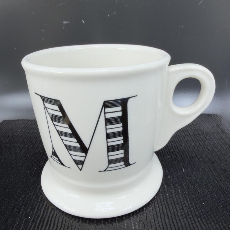 May include: White ceramic mug with a black, striped letter "M" on the front. The mug has a rounded base and a handle. The mug is on a dark surface.