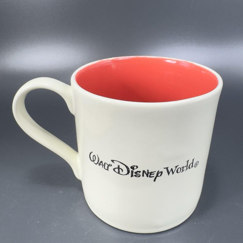 May include: A white ceramic mug with a red interior. The mug has a curved handle and the words "Walt Disney World" are printed in black script on the side. The mug is on a gray surface.