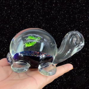 May include: A clear glass turtle figurine with a round, dome-shaped shell containing two green frogs. The turtle's head and legs are also made of clear glass.