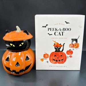 May include: A ceramic lidded mug shaped like a black cat emerging from an orange jack-o'-lantern, accompanied by its white product box labeled PEEK-A-BOO CAT with illustrations of cats and pumpkins and the Pottery Barn brand name printed below.