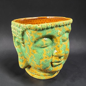May include: A ceramic Buddha head planter in shades of green and yellow. The planter has a textured surface and a wide opening. The Buddha's face is serene, with closed eyes and a gentle smile. The planter is a unique decorative item.