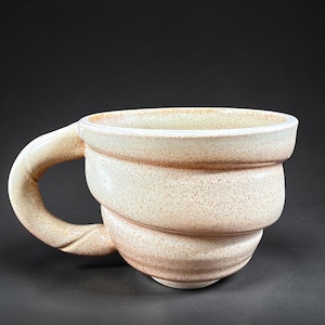 May include: A ceramic mug with a speckled cream and light brown glaze. The mug features a wide, rounded body with a thick, curved handle attached to the side. The surface has a textured, earthy appearance with subtle variations in color tone.
