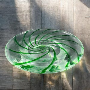May include: Oval glass bowl with a green spiral design. The clear glass bowl has a swirl of green lines that create a spiral pattern. The bowl is sitting on a wooden surface.