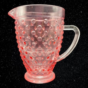 May include: A pink glass pitcher with a textured, dotted surface. The pitcher has a handle and a wide mouth.