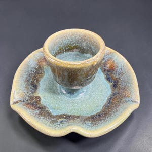 May include: A ceramic cup and matching saucer set featuring a reactive glaze with a blend of light blue, teal, and earthy brown tones. The saucer has a distinct three-lobed, petal-like shape, while the cup has a rounded, deep bowl design.
