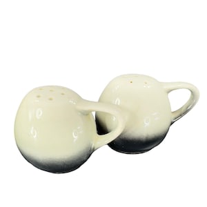 May include: A set of two white ceramic salt and pepper shakers with black accents and a handle. The shakers have a round shape and a gradient design.