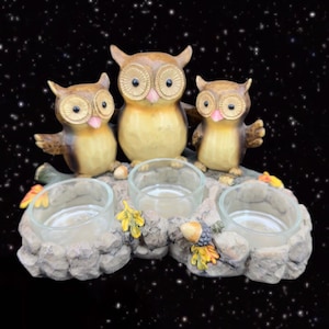 May include: A three-piece owl candle holder set with three glass candle holders. The owls are brown, beige, and white with black eyes and pink beaks. The candle holder is made of a stone-like material and has a rustic look.