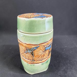 May include: A ceramic jar with a lid. The jar is light green with a band of brown and tan leaf patterns around the middle. The lid has a similar leaf pattern with blue accents. The jar is cylindrical and has a smooth, glossy finish.