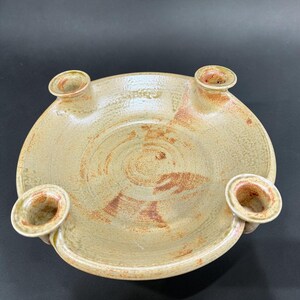 May include: A round, beige ceramic candle holder with a textured, speckled surface and reddish-brown accents. The piece features a shallow, bowl-like center and four evenly spaced, raised circular candle sockets integrated into the outer rim.