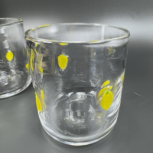 May include: Two clear drinking glasses with yellow lemon designs. The glasses are cylindrical with a slightly textured surface. The yellow lemon designs are scattered around the glass, adding a touch of whimsy. Perfect for serving beverages.
