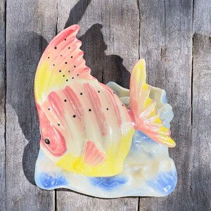 May include: A ceramic wall planter shaped like a pink and yellow fish with black spots. The fish is swimming in a blue and white wave design.