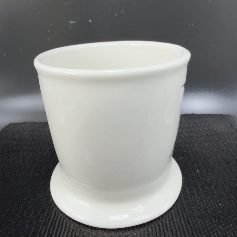May include: A white ceramic shaving mug with a rounded base and a slightly flared top. The mug has a smooth, glossy finish and appears to be empty. The mug is designed to hold shaving soap and create a rich lather.