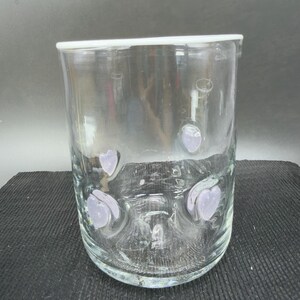 May include: A clear glass vase with a white rim and lavender heart-shaped accents. The vase is cylindrical and appears to be made of thick glass. The heart accents are evenly spaced around the lower portion of the vase.