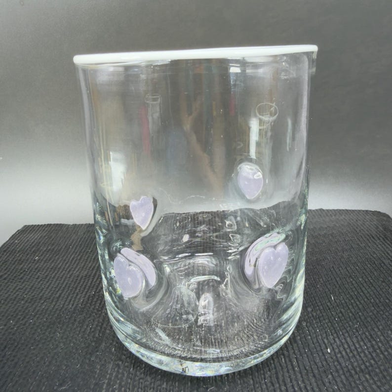 May include: A clear glass vase with a white rim and lavender heart-shaped accents. The vase is cylindrical and appears to be made of thick glass. The heart accents are evenly spaced around the lower portion of the vase.