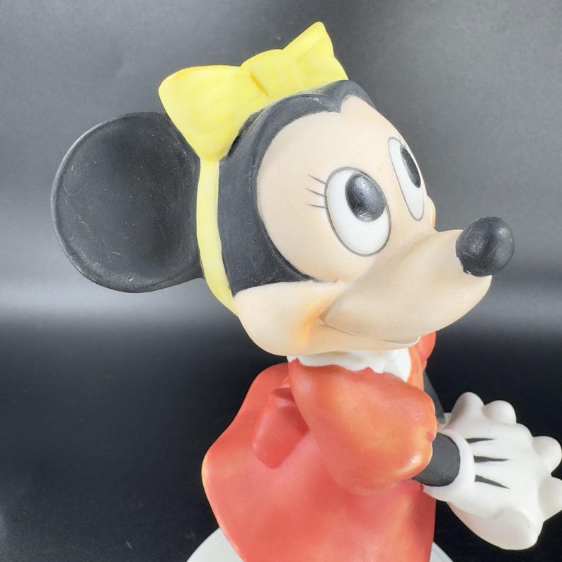 May include: A porcelain figurine of Minnie Mouse wearing a red dress and a yellow bow. She has black ears and a white face with black eyes.