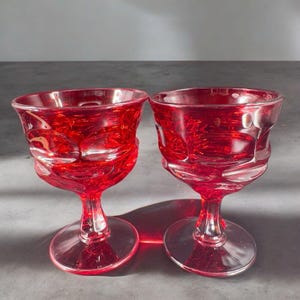 May include: Two red glass goblets with a textured design. The goblets have a round bowl and a tall stem with a wide base.