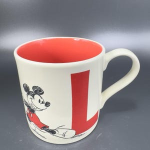 May include: Cream-colored ceramic mug with a red interior and a large red letter "L". The mug features a black and red illustration of Mickey Mouse. The handle is white.