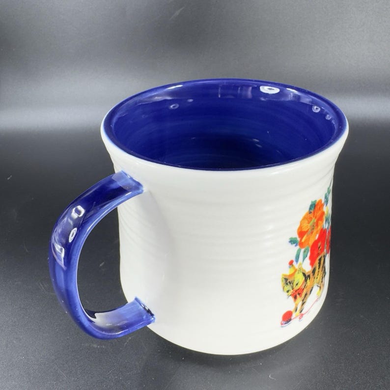 Puede incluir: Taza de cer&aacute;mica blanca con interior y asa azules. La taza presenta un dise&ntilde;o floral con flores naranjas y rojas y un gato de dibujos animados. El asa y el interior son de color azul oscuro.