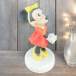 May include: A white ceramic figurine of a cartoon mouse wearing a red dress and yellow shoes. The mouse has black ears and a yellow bow. The figurine is standing on a white base.