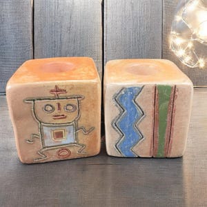 May include: Two square, ceramic candle holders. One is decorated with a figure, the other with a blue zigzag and green and brown stripes. Both have a circular indentation at the top for a candle. The candle holders are tan and orange.