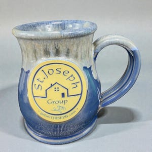 May include: A handmade ceramic mug with a blue and gray glaze. The mug features a large handle and a yellow circular logo with the text "St. Joseph Group" and a house graphic. The website address "gotoSTJOSEPH.com" is also visible.