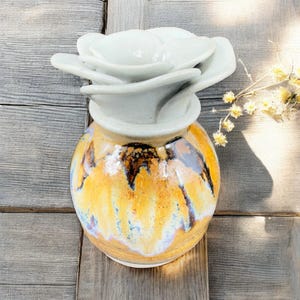 May include: A ceramic vase with a white, flower-shaped top and a round, glazed body. The body features a blend of yellow, orange, and brown hues with blue accents. The vase is a decorative item.