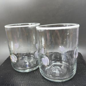 May include: Two clear drinking glasses with a white rim and lavender heart-shaped designs. The glasses are cylindrical and appear to be made of glass. The glasses are sitting on a dark surface.