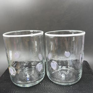 May include: Two clear glass tumblers with a white rim and lavender heart-shaped designs. The glasses are cylindrical and appear to be handmade. The background is a dark grey.