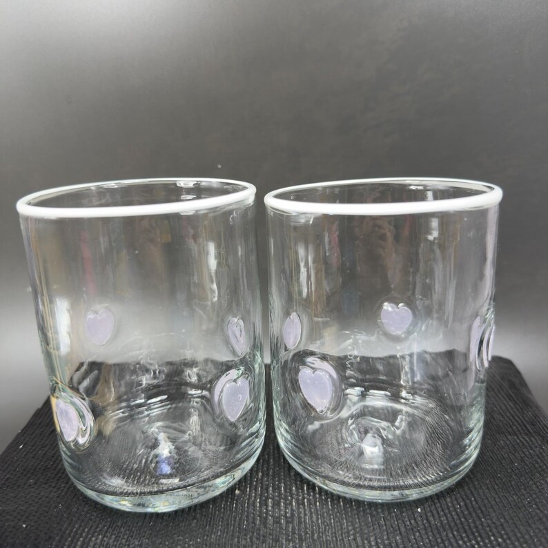 May include: Two clear glass tumblers with a white rim and lavender heart-shaped designs. The glasses are cylindrical and appear to be handmade. The background is a dark grey.