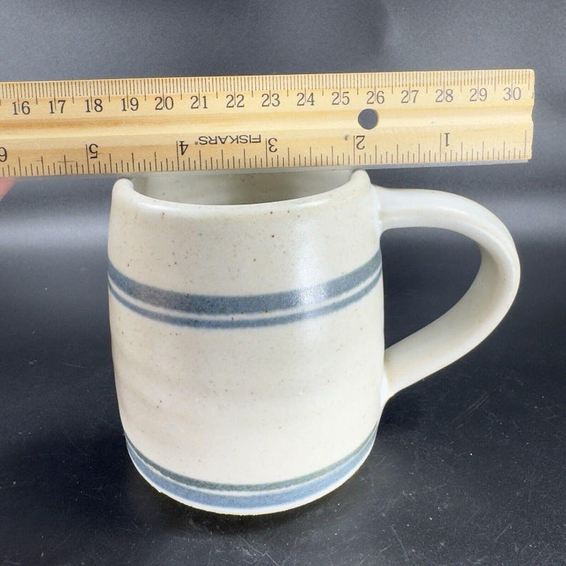 May include: Cream-colored ceramic mug with two blue stripes and a handle. The mug is approximately 4 inches tall and is being measured with a wooden ruler. The mug has a speckled texture.