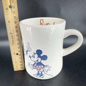 May include: White ceramic coffee mug with a blue and red illustration of Mickey Mouse and Minnie Mouse. The mug has the word "Fun!" written in red on the front.