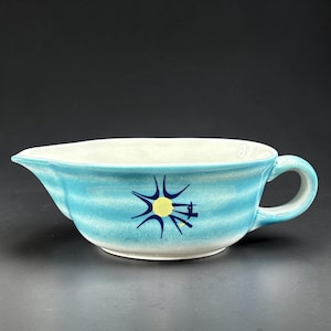 May include: A ceramic gravy boat featuring a light blue and white horizontal gradient glaze. The exterior is decorated with a central dark blue star-like graphic with a yellow circular center. It has a curved handle and a pouring spout.