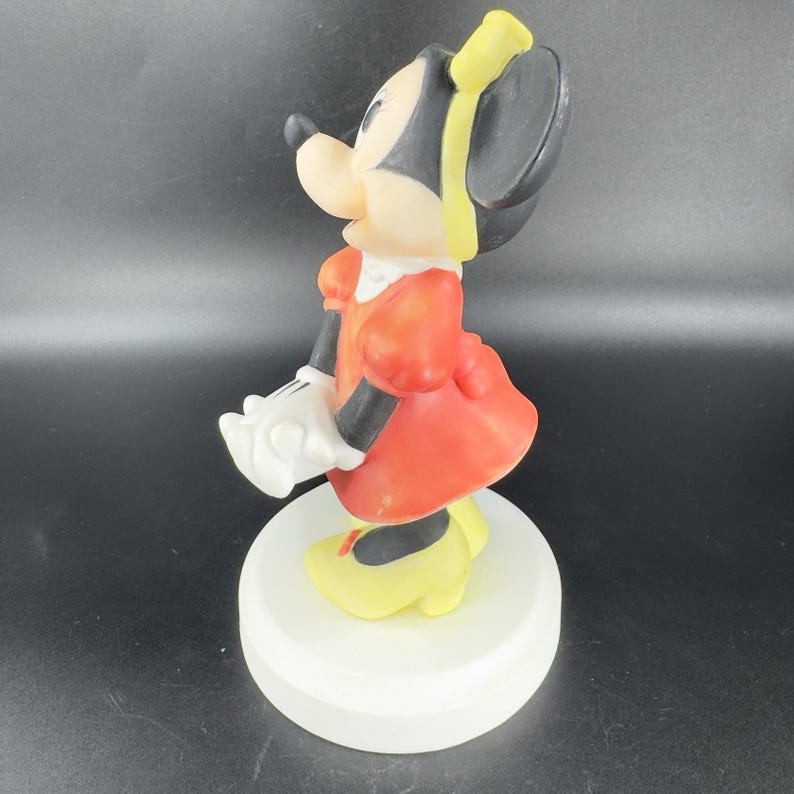 May include: A red and white figurine of Minnie Mouse wearing a yellow bow. She is standing on a white base.