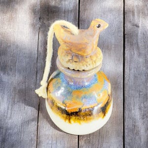 May include: A ceramic bottle with a bird-shaped stopper and a string attached. The bottle is decorated with a colorful glaze in shades of blue, yellow, and brown.