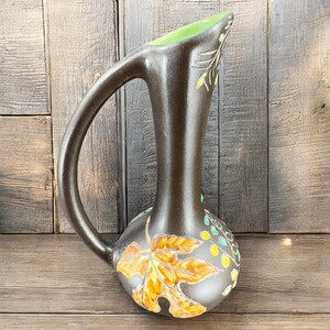 Puede incluir: Una jarra de cerámica marrón con la parte superior verde y un asa. La jarra está decorada con hojas y bayas pintadas en tonos naranja, amarillo y verde.