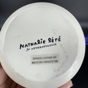 Puede incluir: Plato de cer&aacute;mica blanco con el texto "NATHALIE R&Eacute;T&Eacute; for ANTHROPOLOGIE". El plato est&aacute; marcado con "DISHWASHER & MICROWAVE SAFE" y "MADE IN CHINA / FABRIQU&Eacute; EN CHINE".
