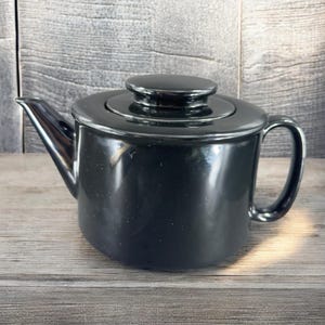 May include: A black ceramic teapot with a lid and a handle. The teapot is sitting on a wooden surface.