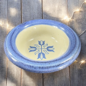 May include: A handmade ceramic bowl with a light yellow interior and a blue rim. The bowl features a blue floral design in the center. The bowl is oval-shaped and sits on a wooden surface.
