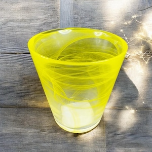 May include: A bright yellow glass vase with a flared top. The vase has a translucent quality with swirling white patterns throughout. The vase is sitting on a weathered wooden surface.