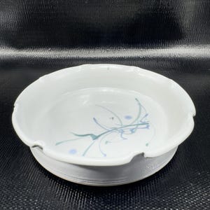 May include: White ceramic serving dish with a scalloped rim and a pedestal base. The interior features abstract blue and green brushstrokes. The dish is round and appears to be handmade.