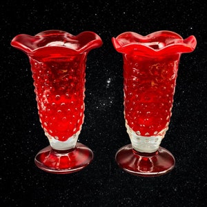 May include: Two red glass vases with a textured, bubble-like surface. The vases have a flared top and a round base.