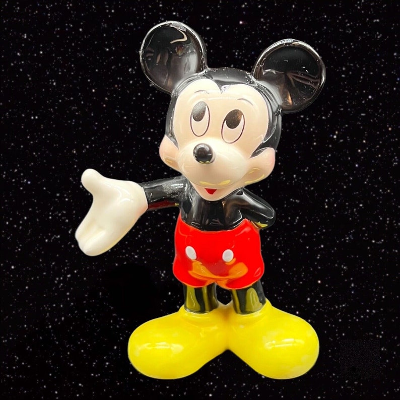 Puede incluir: Una figurita de cer&aacute;mica de Mickey Mouse, de pie con el brazo derecho extendido y la mano izquierda en la cadera. Lleva sus caracter&iacute;sticos pantalones cortos rojos con botones blancos y zapatos amarillos. La figurita est&aacute; sobre un fondo negro con estrellas blancas.