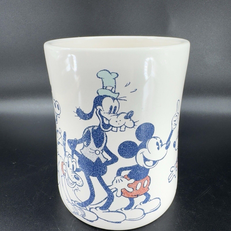 May include: White ceramic mug with a blue and white illustration of Mickey Mouse, Goofy, and Pluto. The characters are in a cartoon style and are smiling.