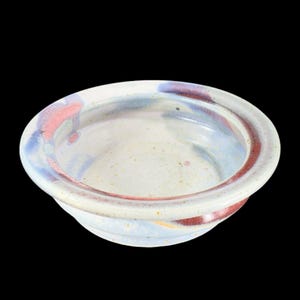 May include: A white ceramic bowl with a light blue and pink glaze. The bowl has a wide rim and a slightly rounded bottom.