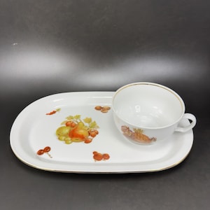 May include: White ceramic teacup and saucer set. The oval saucer and cup feature a gold rim and a fruit design with pears, grapes, and berries. The cup has a handle and a matching fruit design.