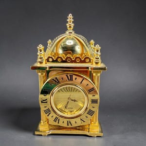 May include: A gold-colored Bulova clock with Roman numerals on the clock face. The clock has a domed top and a rectangular base with decorative columns. The clock is made of metal and has a shiny finish.