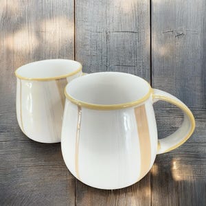 May include: Two white ceramic mugs with a beige and yellow striped design. The mugs have a rounded shape and a handle.