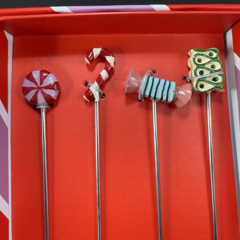May include: A set of four decorative cocktail picks with candy-themed toppers. The toppers include a red and white swirled lollipop, a candy cane, a wrapped candy, and a green and yellow design. The picks are displayed in a red box.