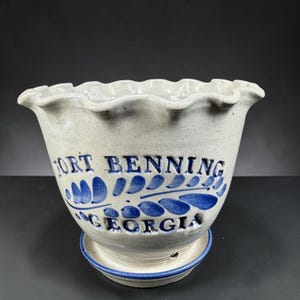 May include: A beige ceramic planter with a ruffled rim and a matching saucer. The planter features blue hand-painted leaf motifs and the embossed text FORT BENNING GEORGIA in blue lettering. The base of the planter includes a small drainage hole.