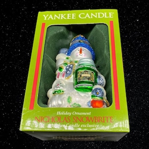 May include: A glass Christmas ornament featuring a snowman holding a Yankee Candle jar. The snowman is wearing a blue hat and scarf and has a white body with green and white accents. The ornament is in a green and red box with the words "Yankee Candle" and "Nicholas Snowbrite" printed on it.